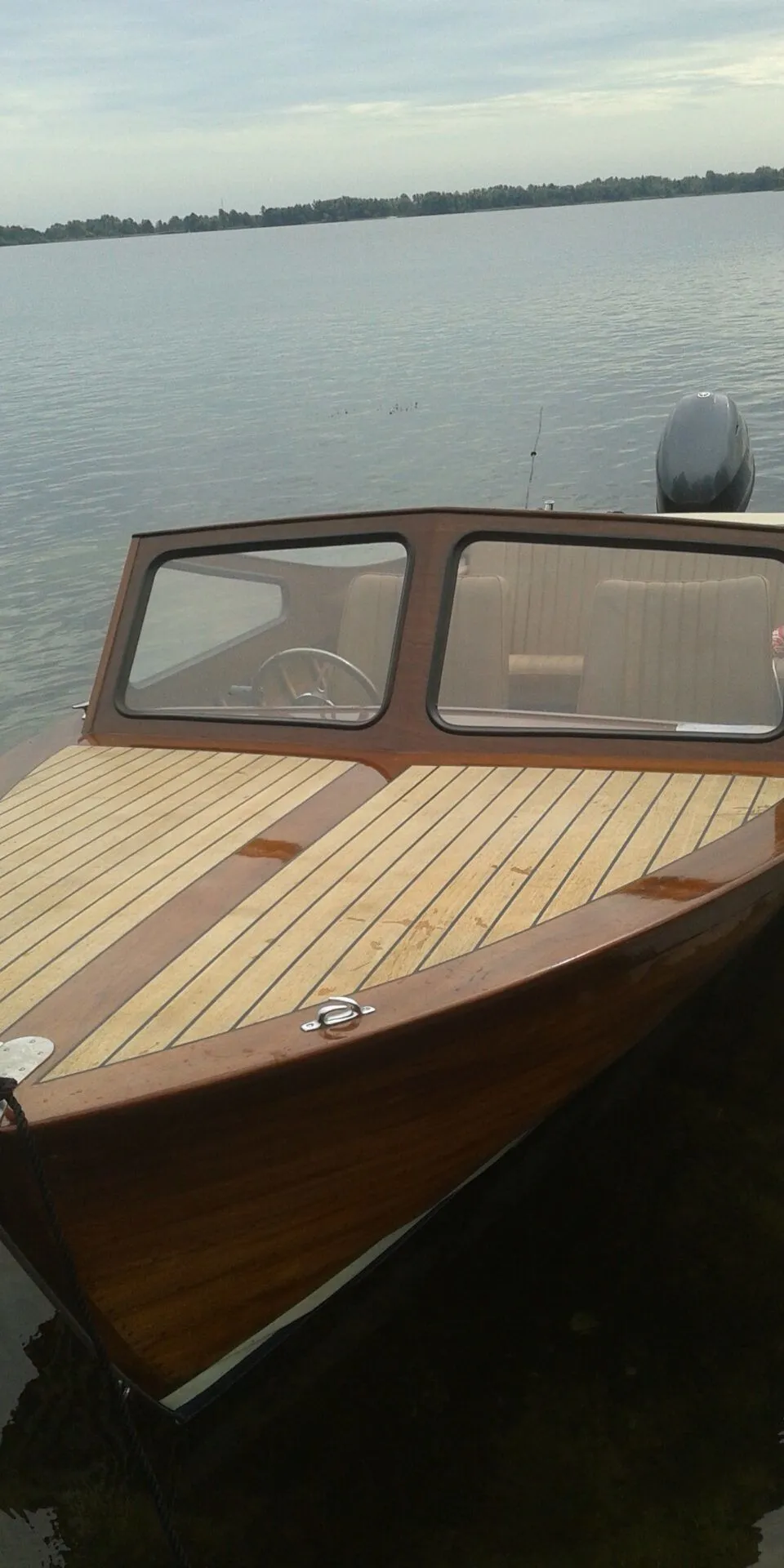 Een houten motorboot met een teakdek en een buitenboordmotor ligt in het water, met een beboste oever op de achtergrond.