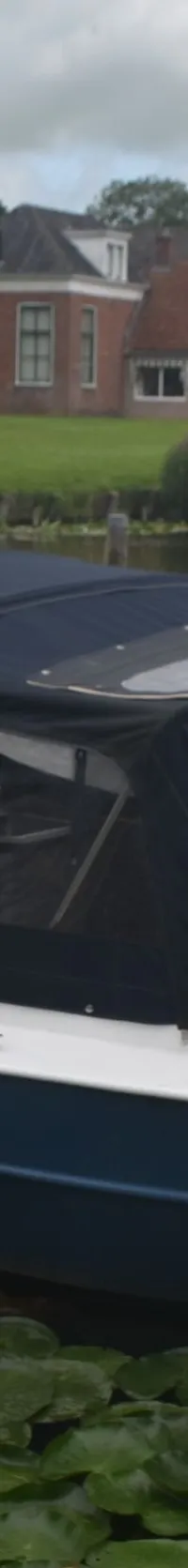 Een witte en blauwe boot met een donkerblauwe kap ligt in het water, omringd door waterlelies, met huizen op de achtergrond.