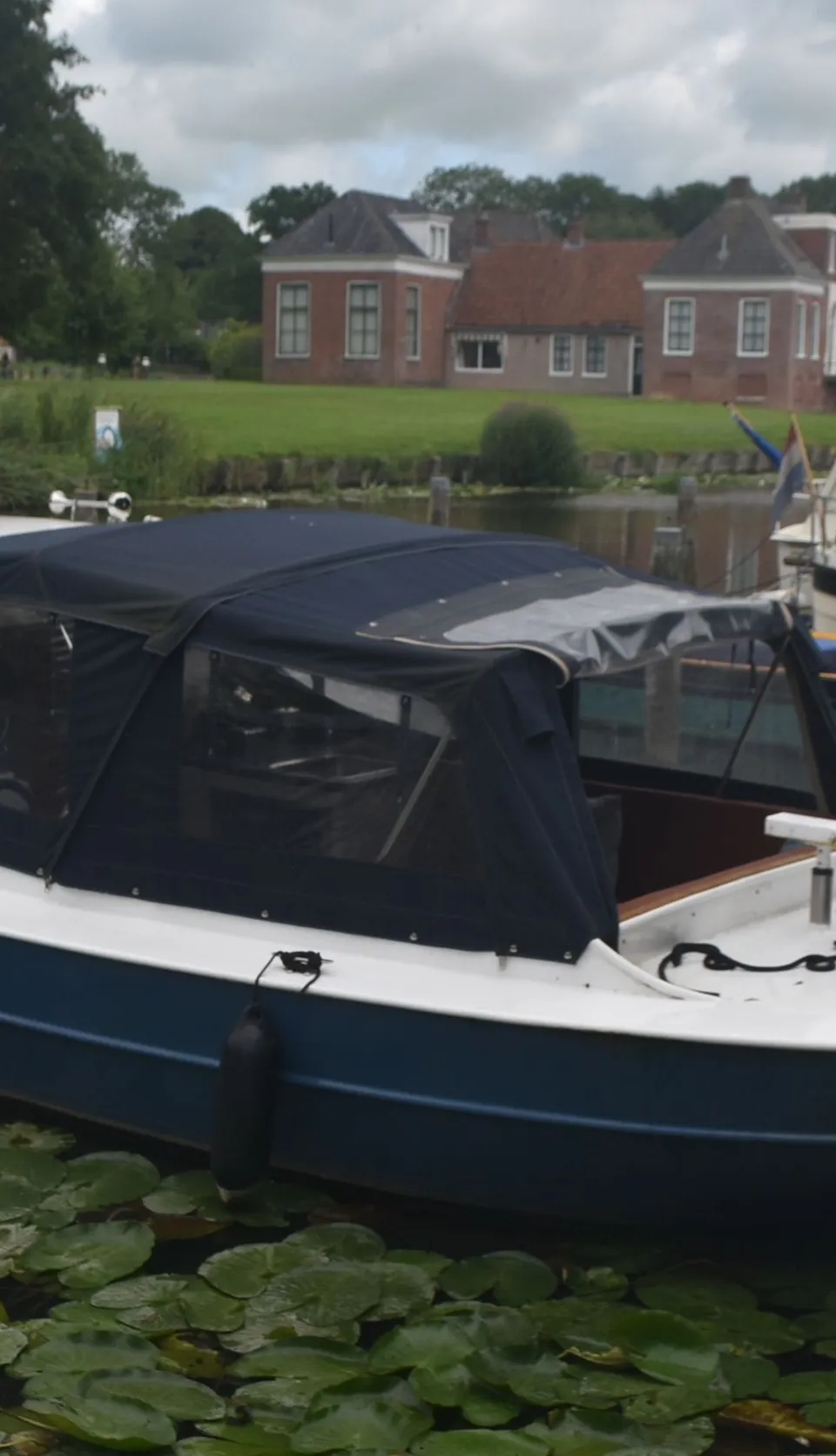 Een witte en blauwe boot met een donkerblauwe kap ligt in het water, omringd door waterlelies, met huizen op de achtergrond.