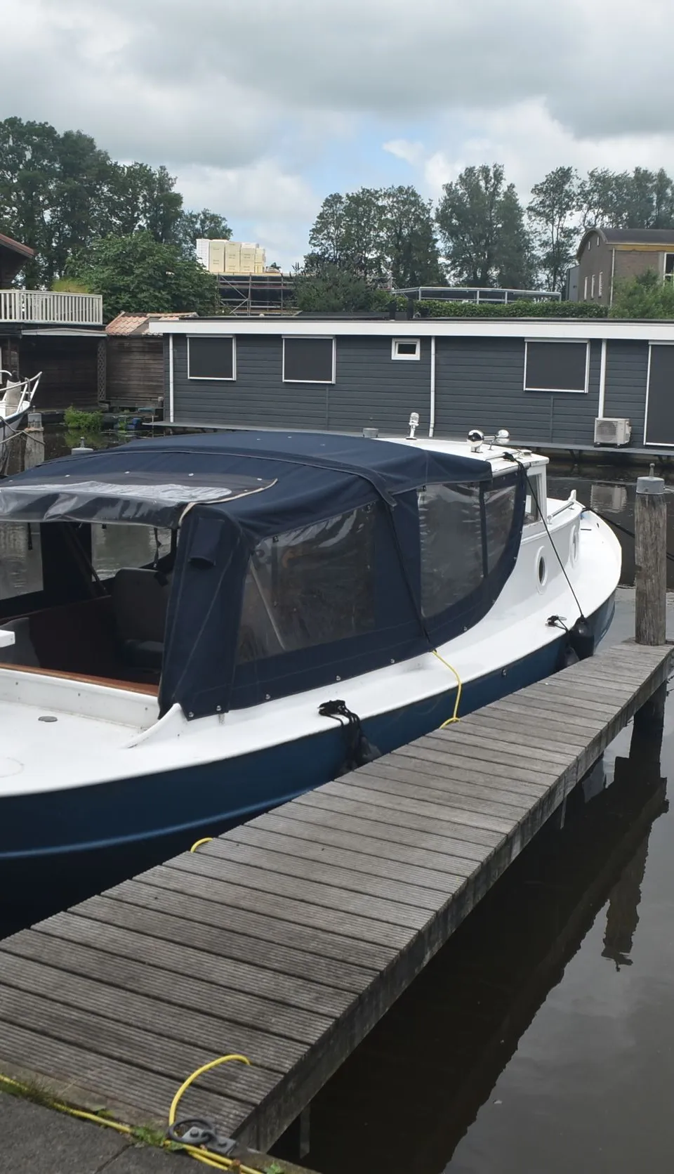 Een witte boot met blauwe romp en donkerblauwe kap ligt afgemeerd aan een houten steiger, omringd door waterlelies.