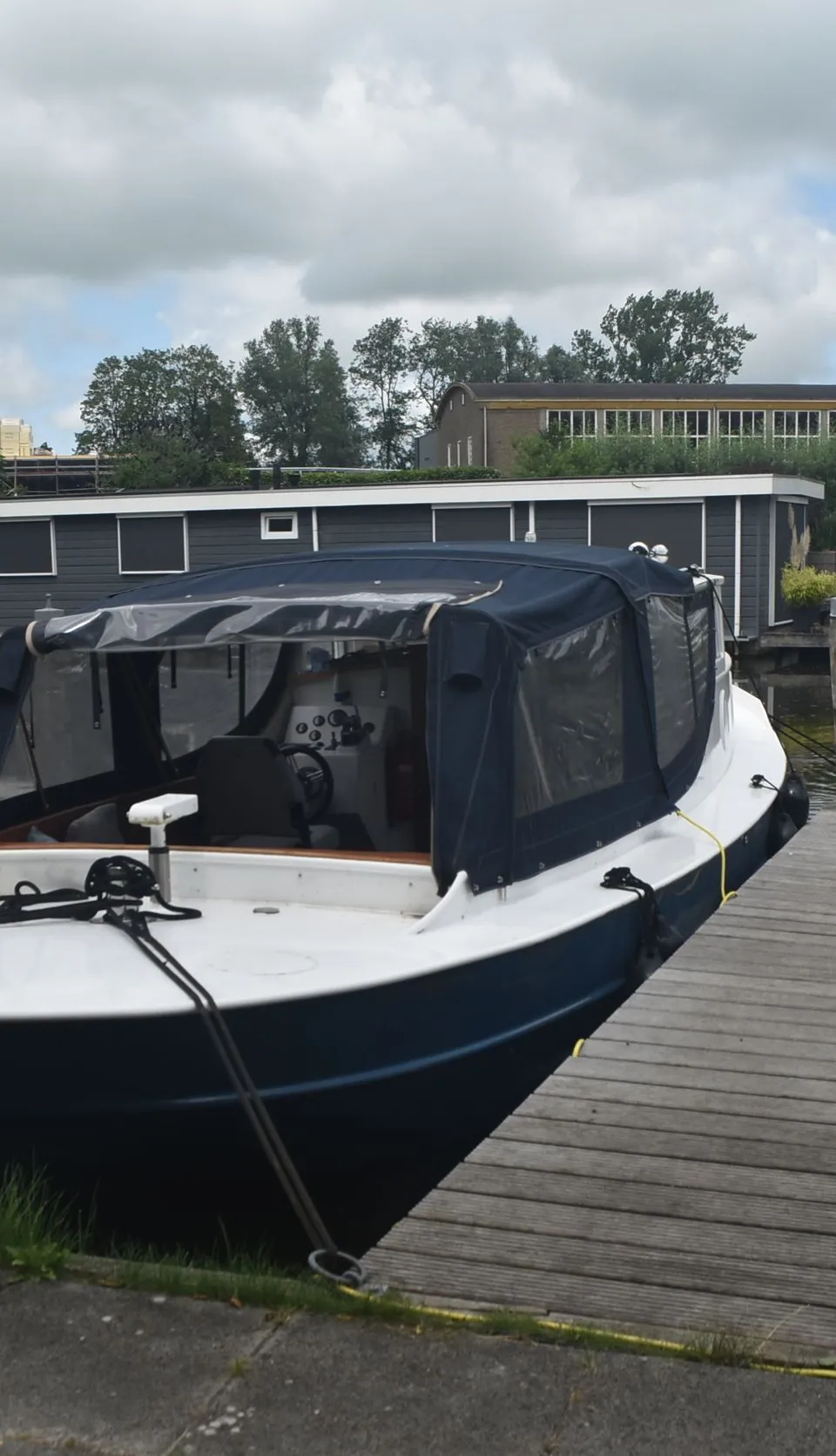 Een witte en blauwe boot met een donkerblauwe kap ligt aangemeerd aan een houten steiger, met waterlelies op het water.