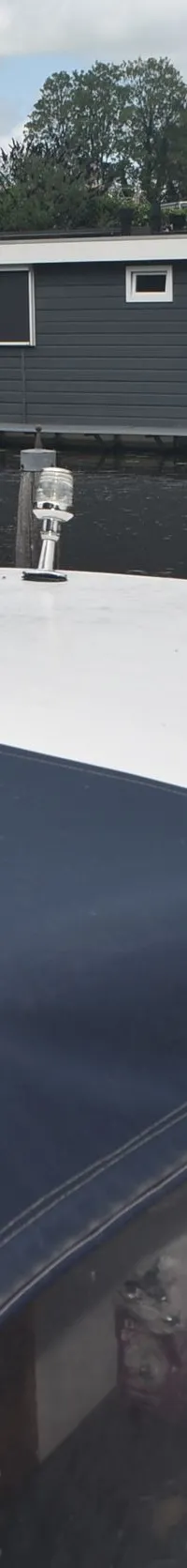 Een witte boot met een donkerblauwe afdekking ligt aangemeerd aan een houten paal, met woonboten op de achtergrond.