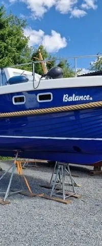 Een blauw-witte motorboot met de naam "Balance" staat op bokken op een grindterrein, met een andere boot ernaast.