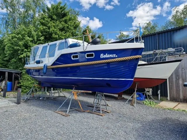 Een blauw-witte motorboot met de naam "Balance" staat op bokken op een grindterrein, met een andere boot ernaast.