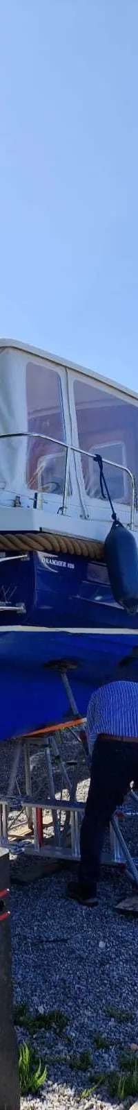 Een blauw-witte motorboot staat op bokken op een grindterrein, met een persoon die aan de onderkant werkt.