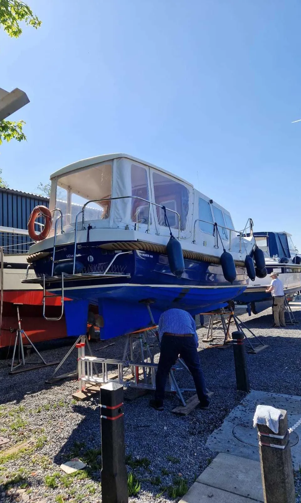 Een blauw-witte motorboot staat op bokken op een grindterrein, met een persoon die aan de onderkant werkt.