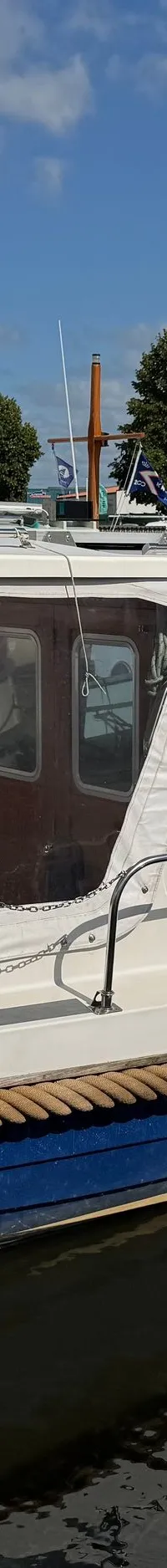 Een witte boot met blauwe romp en een bruine stootrand ligt in het water, met de naam "DRAMMER 820" op de achtersteven.