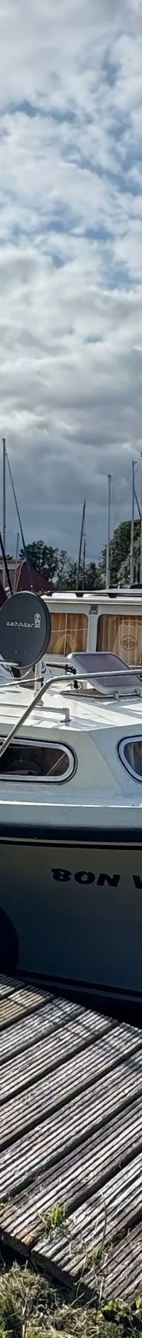 Een witte motorboot met de naam "BON VOYAGE" ligt aangemeerd aan een houten steiger onder een bewolkte hemel.