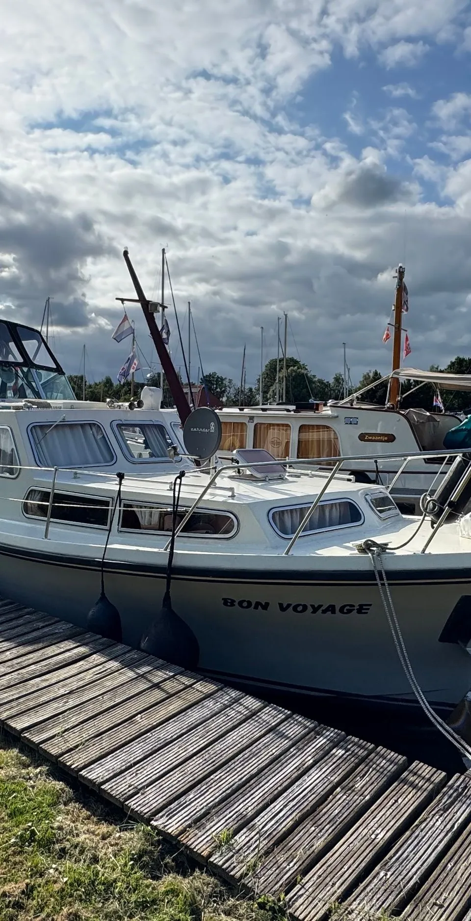 Een witte motorboot met de naam "BON VOYAGE" ligt aangemeerd aan een houten steiger onder een bewolkte hemel.