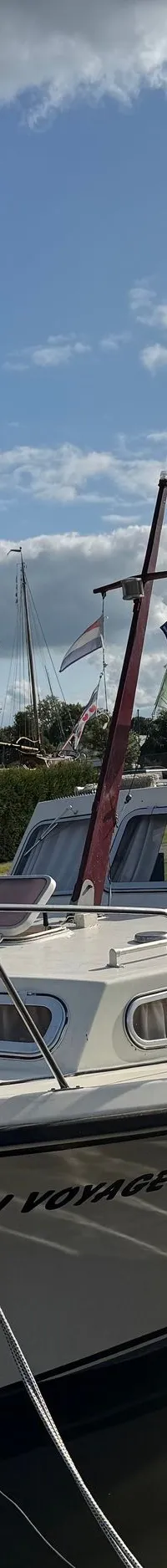 Een witte motorboot met de naam "BON VOYAGE" ligt afgemeerd aan een houten steiger onder een blauwe lucht met wolken.