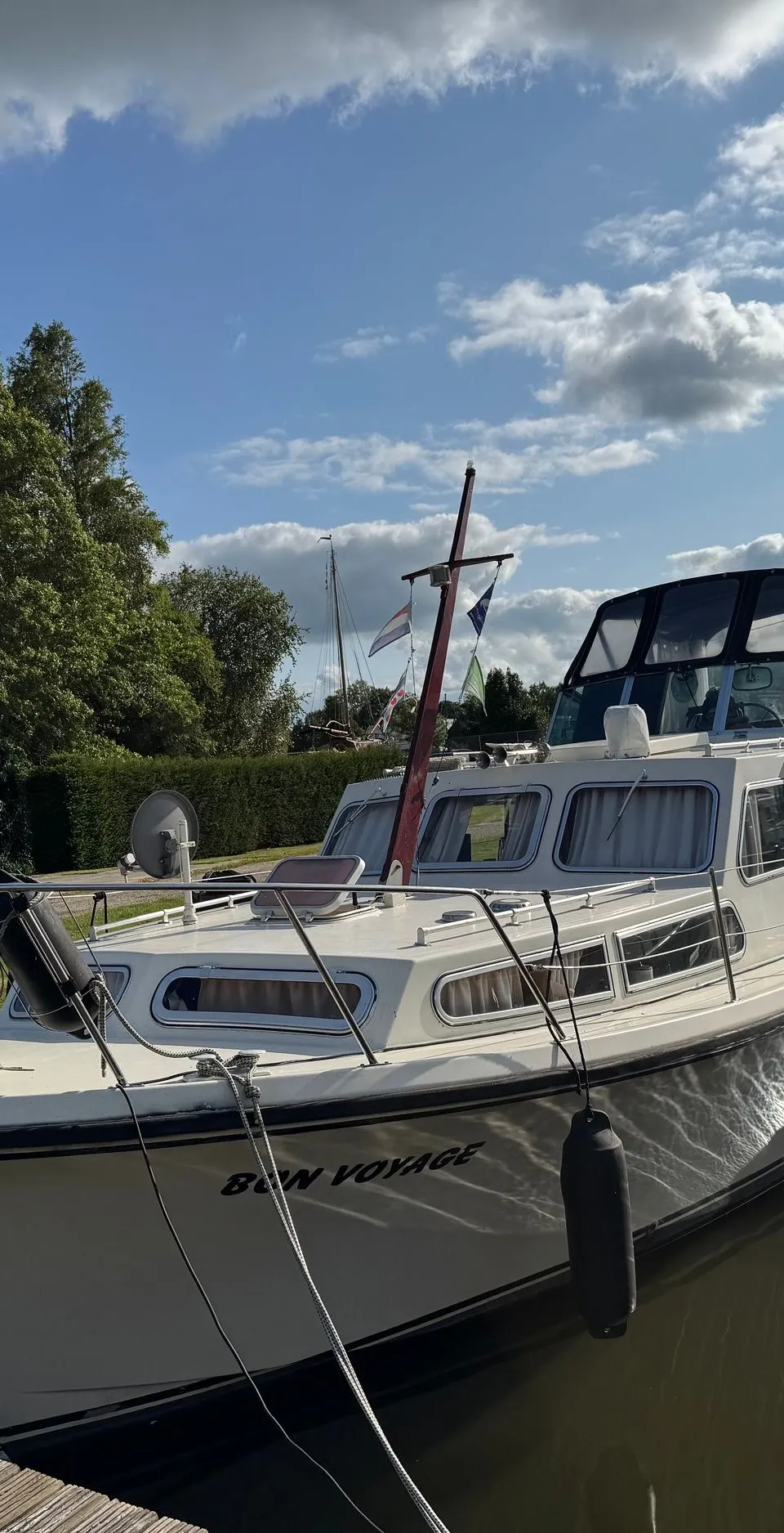 Een witte motorboot met de naam "BON VOYAGE" ligt afgemeerd aan een houten steiger onder een blauwe lucht met wolken.