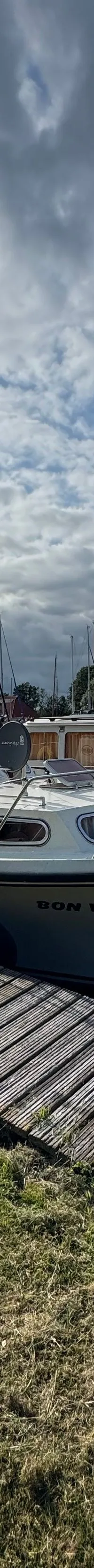 Een witte motorboot met de naam "BON VOYAGE" ligt aangemeerd aan een houten steiger, met gras ernaast. — Neptunus 107 TWIN