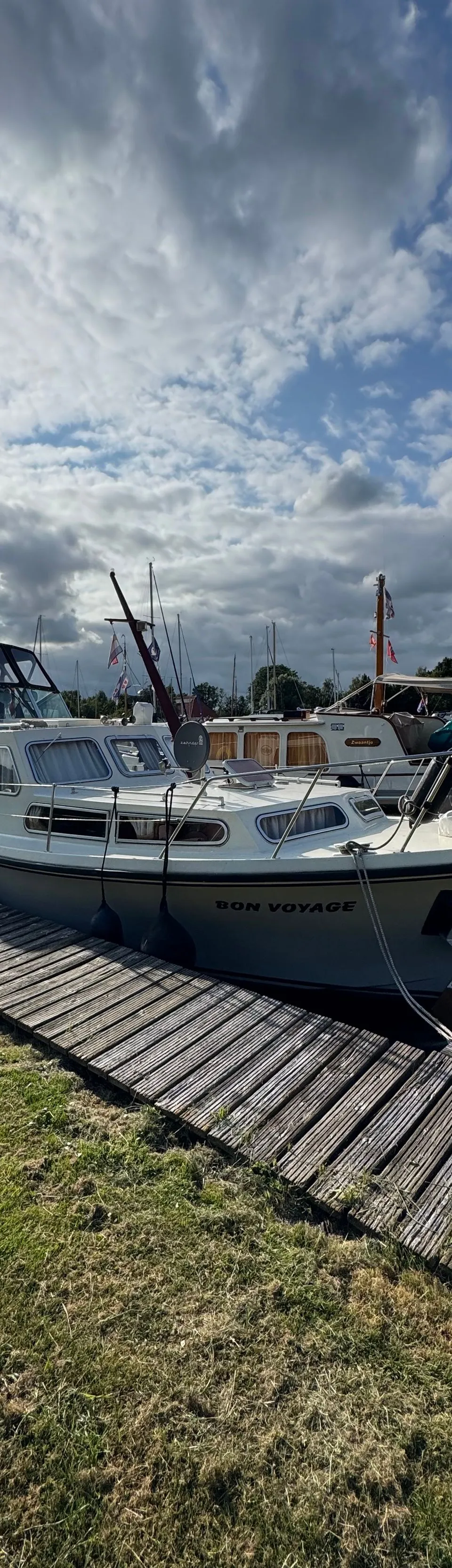 Een witte motorboot met de naam "BON VOYAGE" ligt aangemeerd aan een houten steiger, met gras ernaast. — Neptunus 107 TWIN