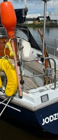 Een blauw-witte zeilboot genaamd "JODZERT" ligt afgemeerd aan een kade, met een oranje boei en gele reddingsboei.