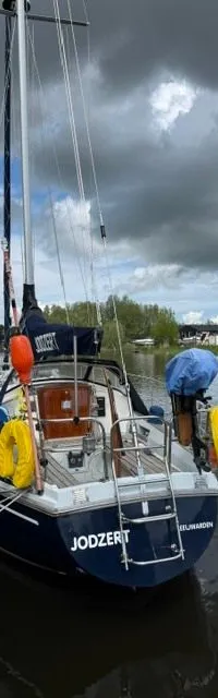 Een blauwe zeilboot met de naam "JODZERT" ligt aangemeerd aan een kade onder een bewolkte hemel. — Victoire 34 S-spant
