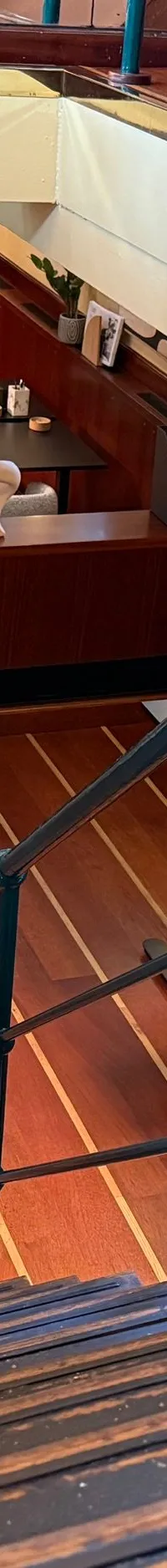 Een houten trap met blauwe reling leidt naar een lager dek met houten vloer, tafels, stoelen en planten.