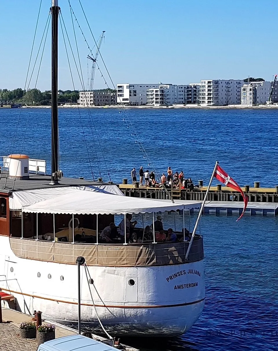 Een grote witte boot met bruine opbouw en twee masten ligt aangemeerd, met "PRINSES JULIANA AMSTERDAM" op de spiegel.