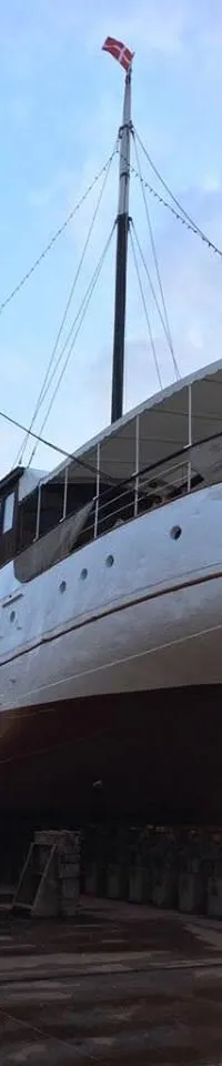 Een witte en bruine boot, genaamd "PRINSES JULIANA AMSTERDAM", staat op houten blokken in een droogdok onder een bewolkte…