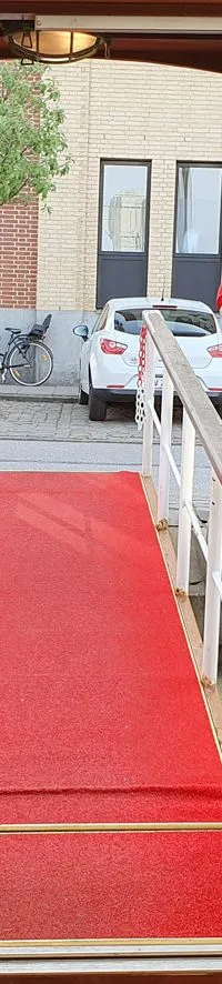 Een rode loper leidt vanaf een houten interieur met tafels en banken naar buiten, geflankeerd door twee Deense vlaggen.