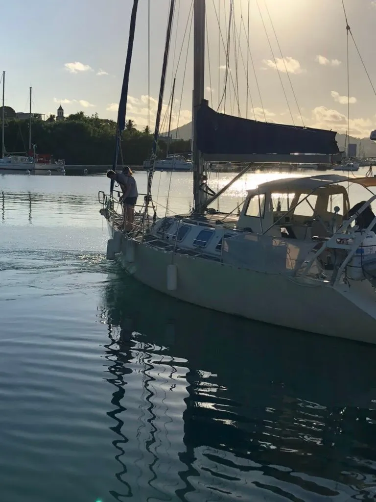 Een zeilboot met een persoon op de boeg ligt in het water, met een deels opgerold grootzeil en fenders aan de zijkant.