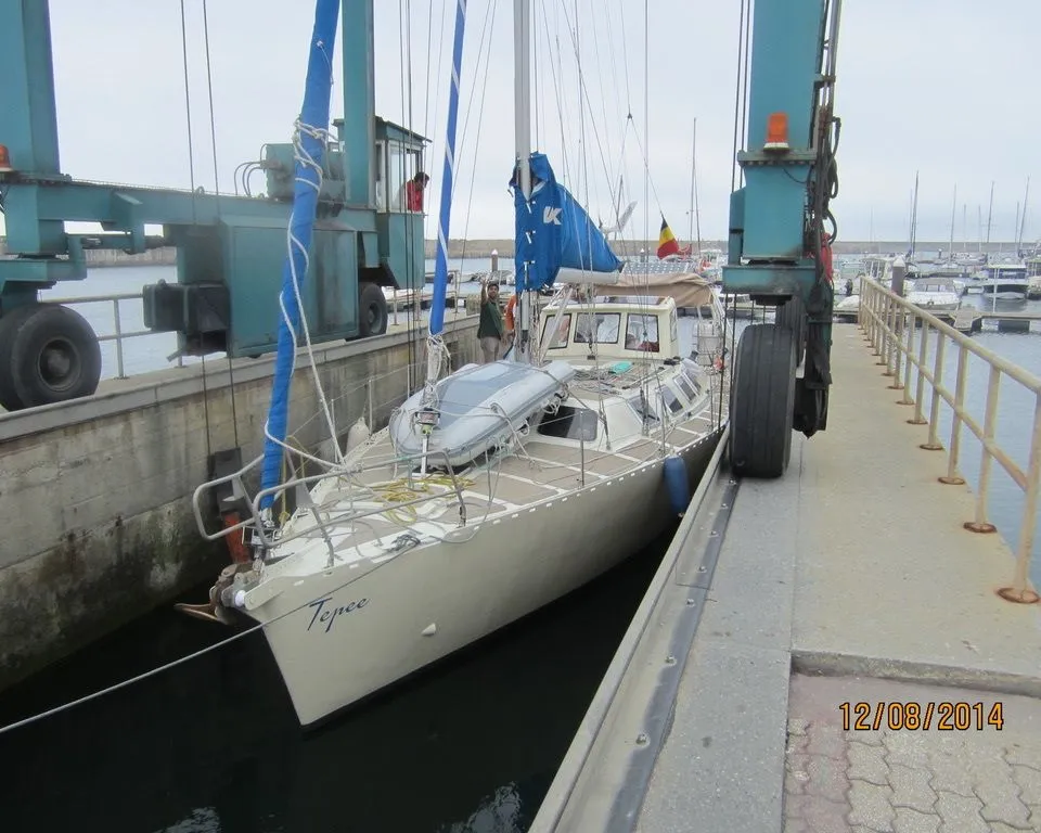 Een lichtgekleurde zeilboot met de naam "Tepee" op de boeg ligt in het water naast een hijskraan in een haven.