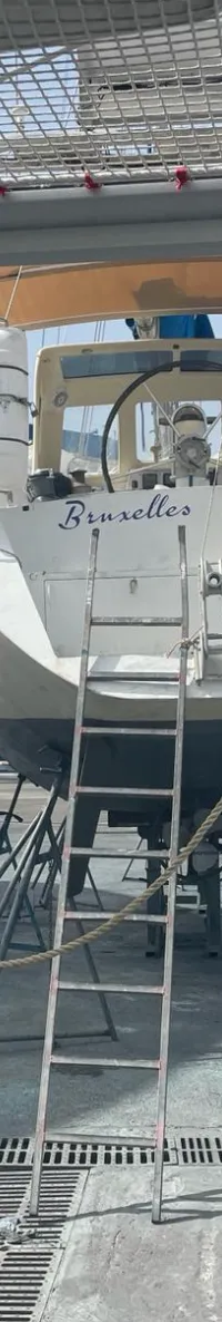 Een witte zeilboot met de naam "Bruxelles" op de spiegel staat op bokken op de wal, met een ladder ertegenaan.