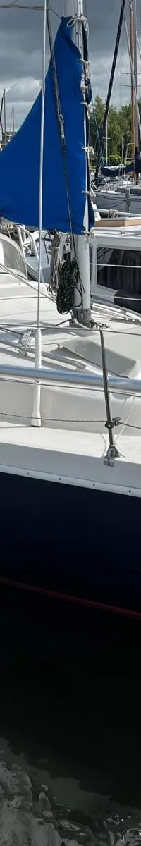 Een zeilboot met een donkerblauwe romp, wit dek, grijze buiskap en een opgerold blauw zeil ligt afgemeerd in een haven.