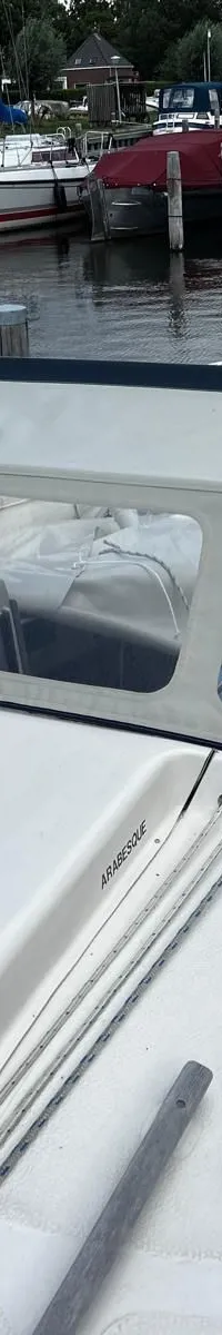 De foto toont het dek van een zeilboot met een blauwe zeilhuik, een lichtgrijze buiskap en de naam "ARABESQUE" op de opbouw.