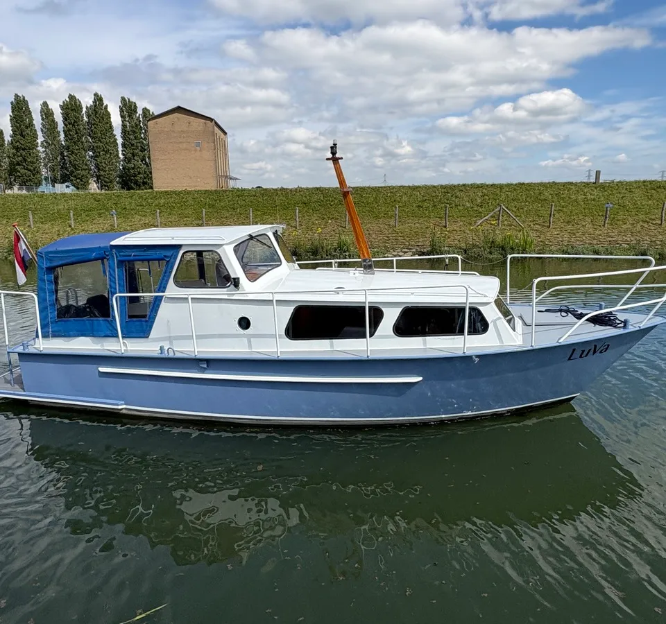 Een blauw-witte motorboot met de naam "LuVa" vaart op een waterweg met een dijk en gebouwen op de achtergrond.