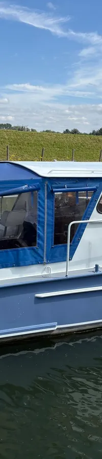 Een blauw-witte motorboot met een blauwe cabriokap vaart op een waterweg met een Nederlandse vlag aan de achtersteven.