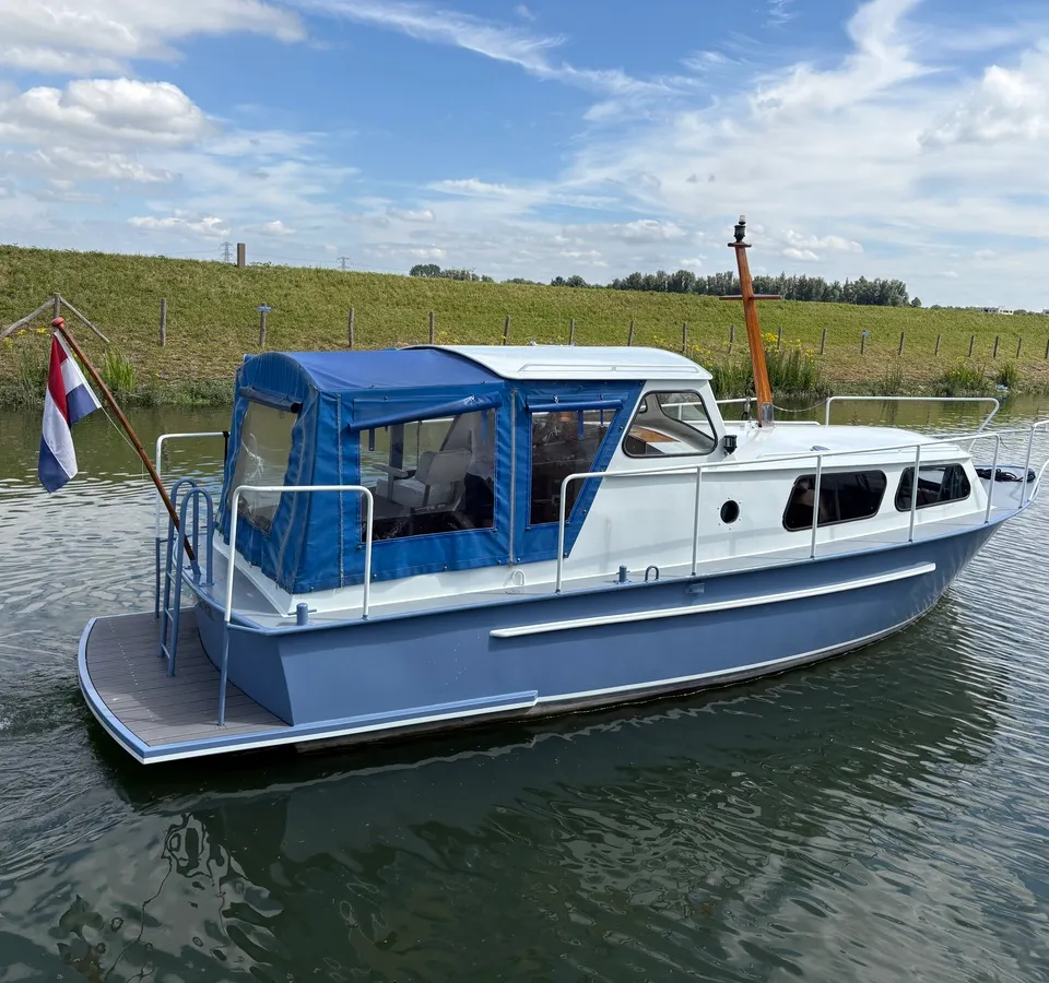 Een blauw-witte motorboot met een blauwe cabriokap vaart op een waterweg met een Nederlandse vlag aan de achtersteven.