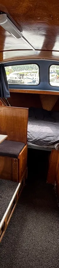 De foto toont het interieur van een boot met houten wanden en plafond, een tafel, een bed en een aanrechtblad.