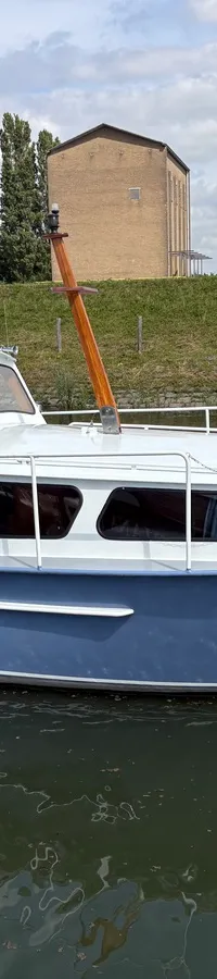 Een blauw-witte motorboot met de naam "LuVa" op de boeg, drijvend op water met een groene oever en gebouwen op de…