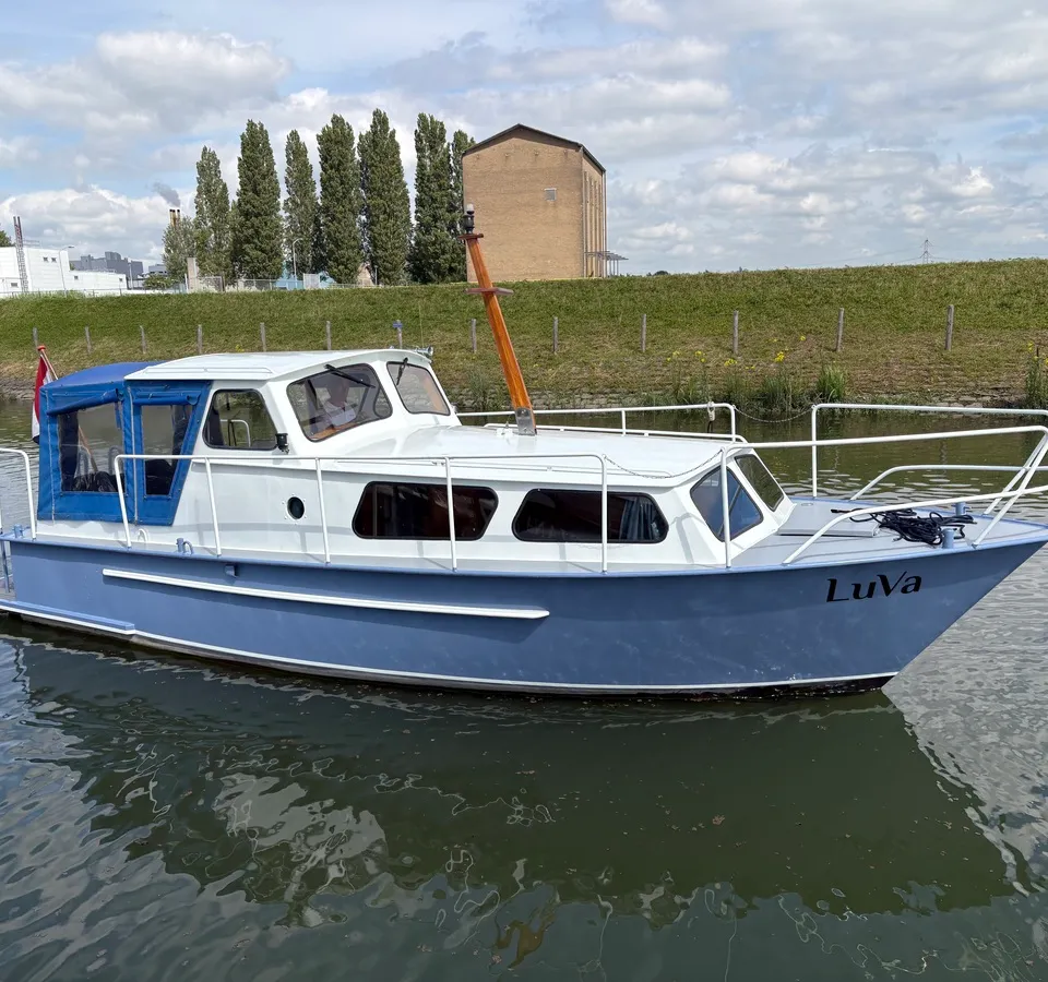 Een blauw-witte motorboot met de naam "LuVa" op de boeg, drijvend op water met een groene oever en gebouwen op de…