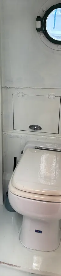 Een witte scheepstoiletruimte met een rond patrijspoortje, een wit toilet en een houten deur met een metalen handvat.