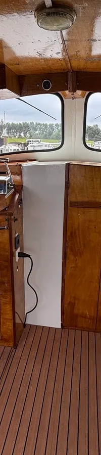 De foto toont het interieur van een stuurhut met een witte stoel, houten stuurwiel en instrumentenpaneel.
