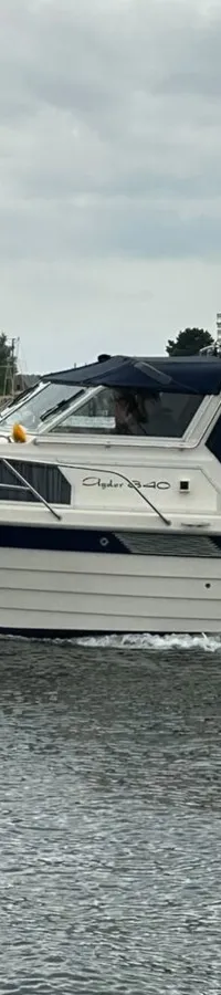 Een witte motorboot met blauwe accenten en een blauwe kap vaart over water met een kade vol zeilboten op de achtergrond.
