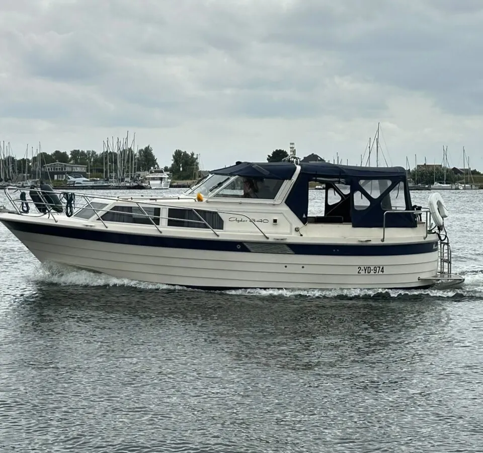 Een witte motorboot met blauwe accenten en een blauwe kap vaart over water met een kade vol zeilboten op de achtergrond.