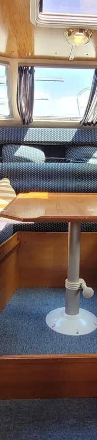 De foto toont een kajuit met houten wanden en plafond, blauwe gestippelde kussens, een houten tafel en ramen met gordijnen.