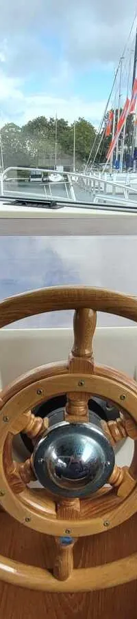 Een houten stuurwiel, een kompas, een Raymarine plotter en diverse meters en schakelaars zijn zichtbaar op het dashboard…