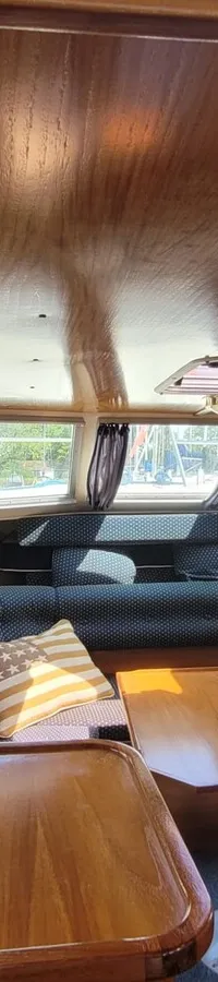 De foto toont het interieur van een boot met houten panelen, ramen met gordijnen en een zithoek met twee tafels.