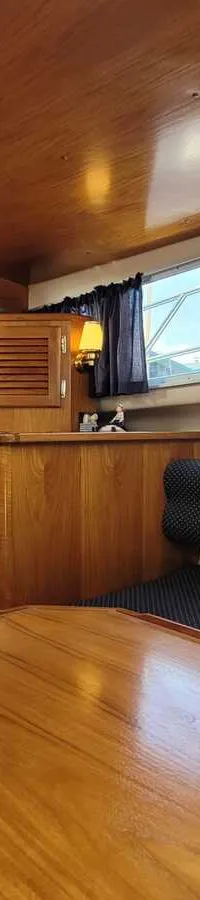 De foto toont het interieur van een boot met houten panelen, een tafel, banken met blauwe kussens en ramen met gordijnen.
