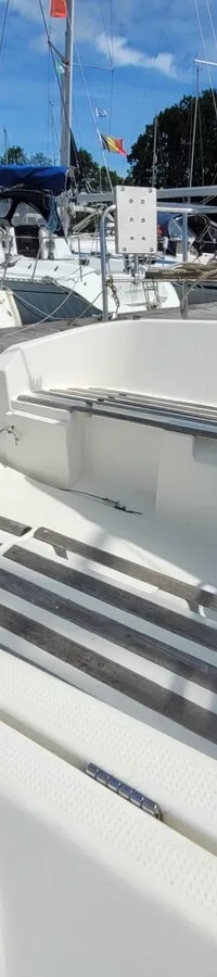 De foto toont het achterdek van een witte zeilboot met een lier, touwen en houten latten, omringd door andere boten in een…