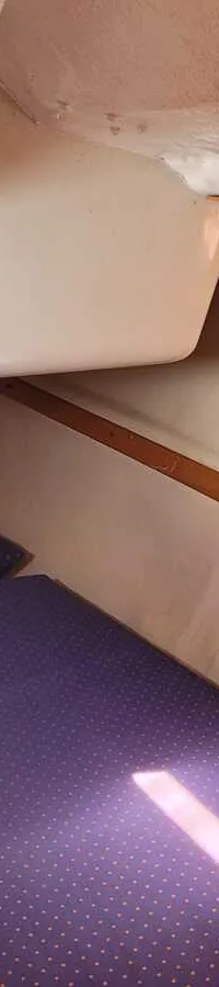 De foto toont een kajuit met twee paarse matrassen, een houten kastje met twee lades en een raam met deels leesbare tekst…