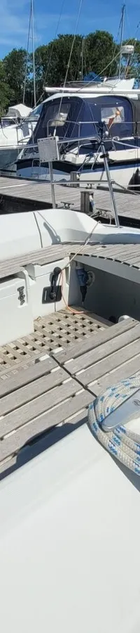 De foto toont de kuip van een witte zeilboot met houten vlonders en touwen, omringd door andere boten in een haven.