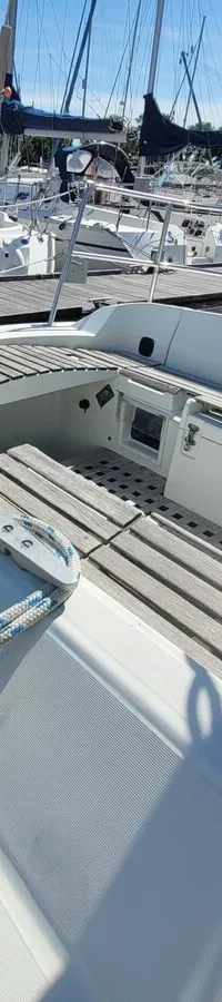 De foto toont het dek en de kuip van een witte zeilboot met houten latten, omringd door andere boten in een haven.