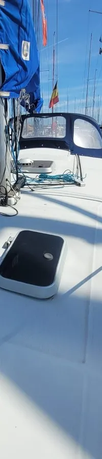 De foto toont het dek van een witte zeilboot met een blauw zeil en een buiskap, omringd door andere boten in een haven.