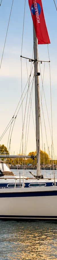Een witte zeilboot met een blauwe streep op de romp ligt op het water, met een rode vlag in de mast en een haven op de…
