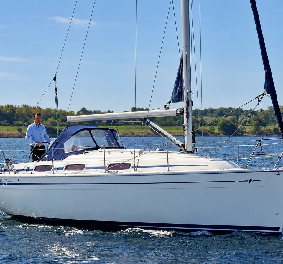 Een witte zeilboot met blauwe strepen en een blauwe buiskap vaart op het water, met een man in een blauw overhemd aan boord.