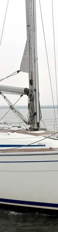 Een witte zeilboot met blauwe strepen en de naam "Diva" op de romp, vaart op rustig water onder een grijze lucht.